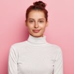 isolated shot pleasant looking young woman wears white turtleneck has hair bun looks with calm facial expressions poses against pink background