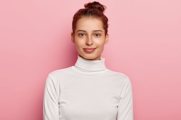 isolated shot pleasant looking young woman wears white turtleneck has hair bun looks with calm facial expressions poses against pink background